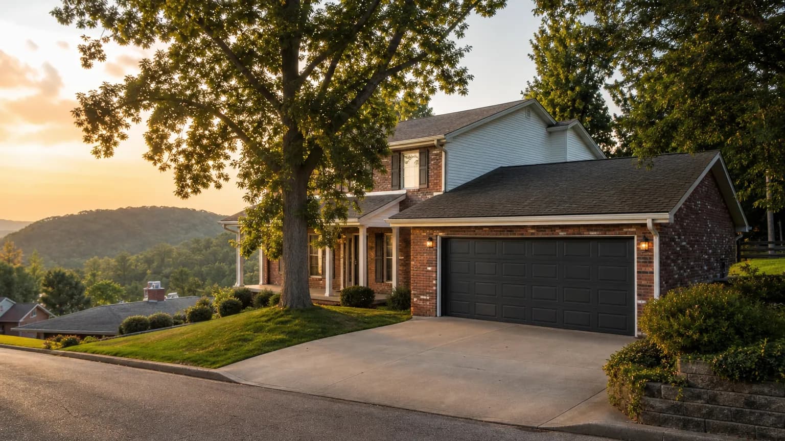 Brick two story home with a white raised panel garage door and a wooded hillside behind it at dusk